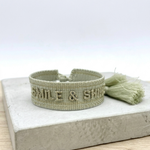 Close-up of adjustable platinum sage woven bracelet with tone-on-tone embroidered “SMILE & SHINE” lettering and sliding knot tassel cords.