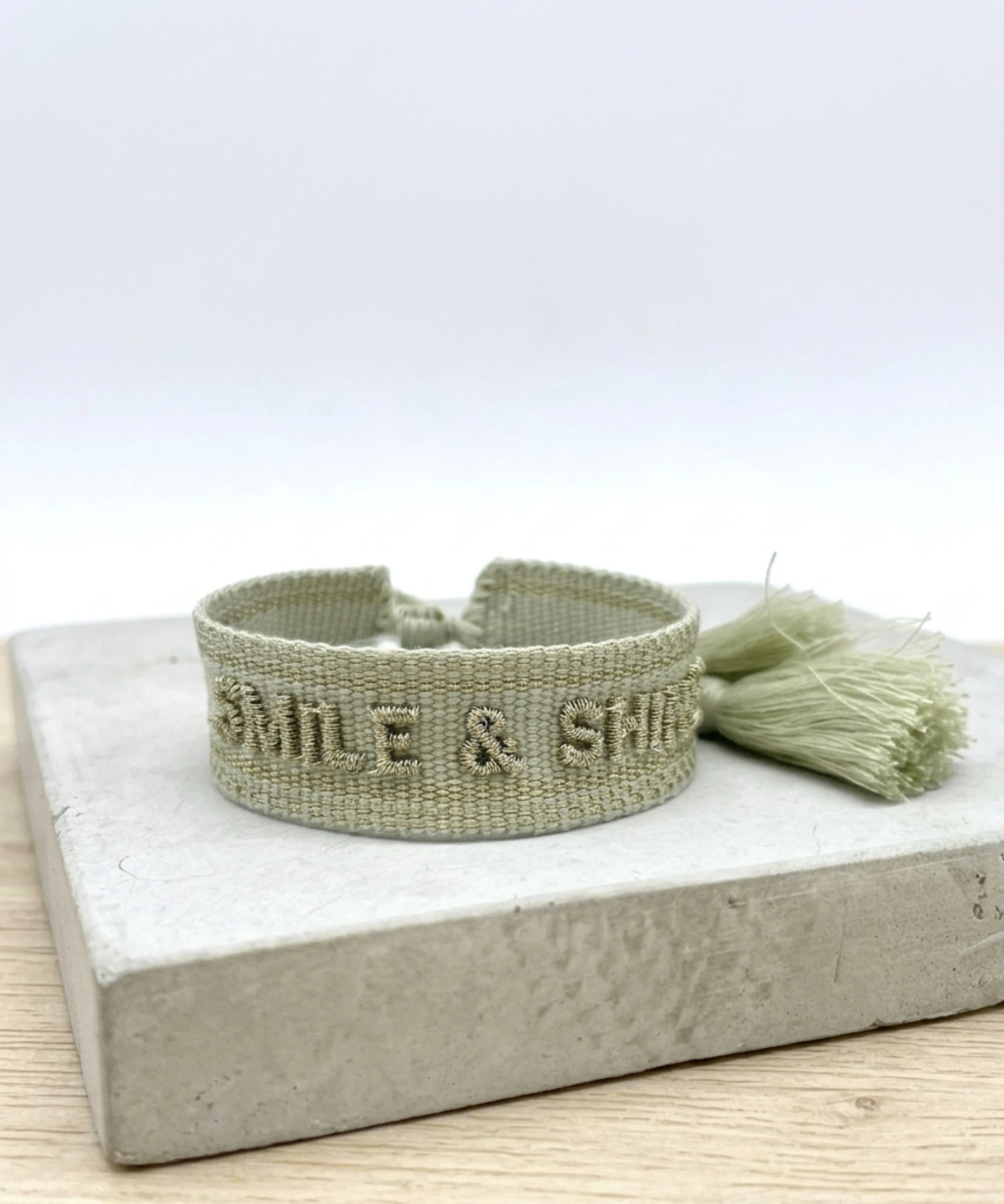 Close-up of adjustable platinum sage woven bracelet with tone-on-tone embroidered “SMILE & SHINE” lettering and sliding knot tassel cords.