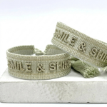 Close-up of adjustable platinum sage woven bracelet with tone-on-tone embroidered “SMILE & SHINE” lettering and sliding knot tassel cords.