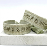 Close-up of adjustable platinum sage woven bracelet with tone-on-tone embroidered “SMILE & SHINE” lettering and sliding knot tassel cords.