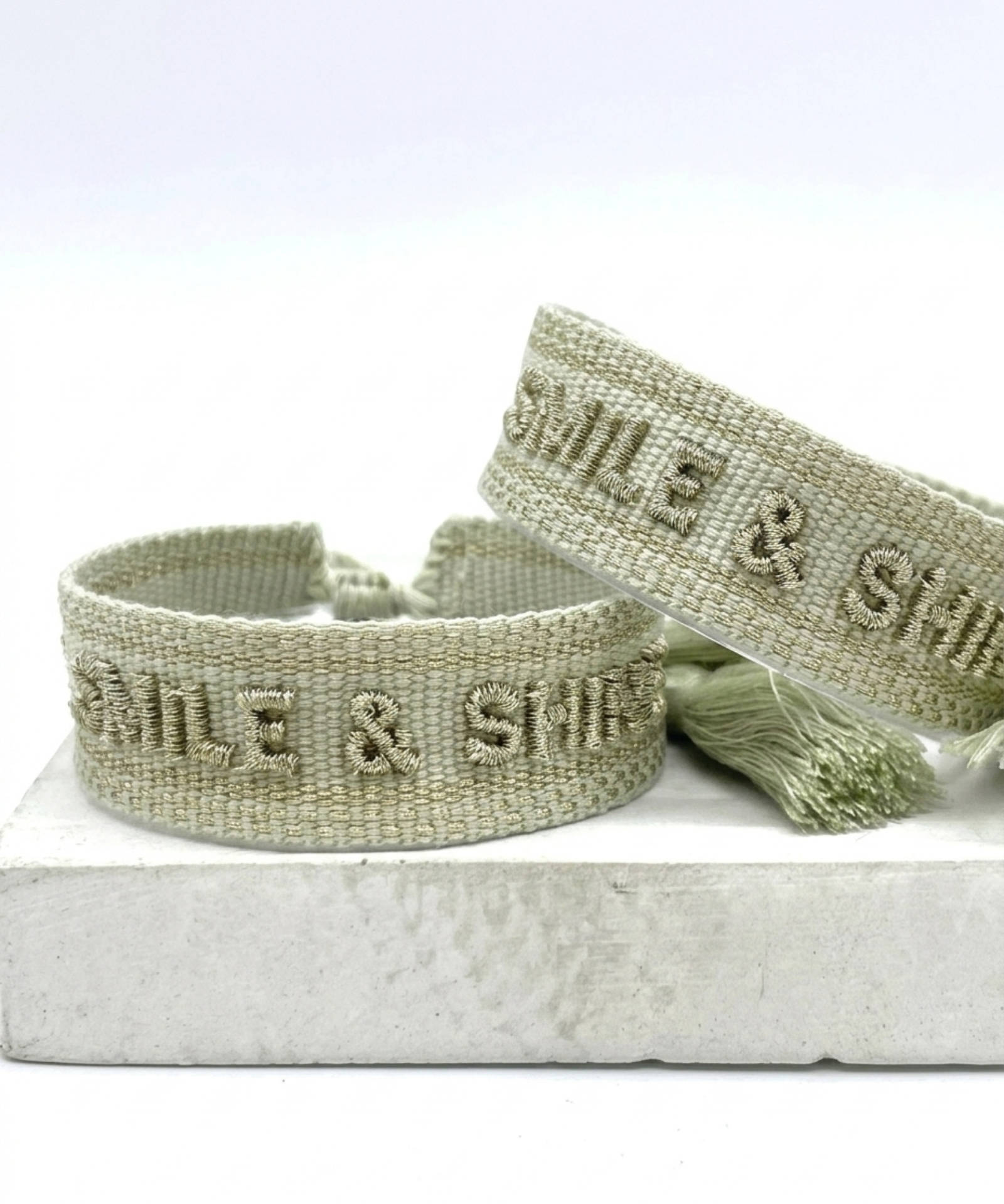 Close-up of adjustable platinum sage woven bracelet with tone-on-tone embroidered “SMILE & SHINE” lettering and sliding knot tassel cords.