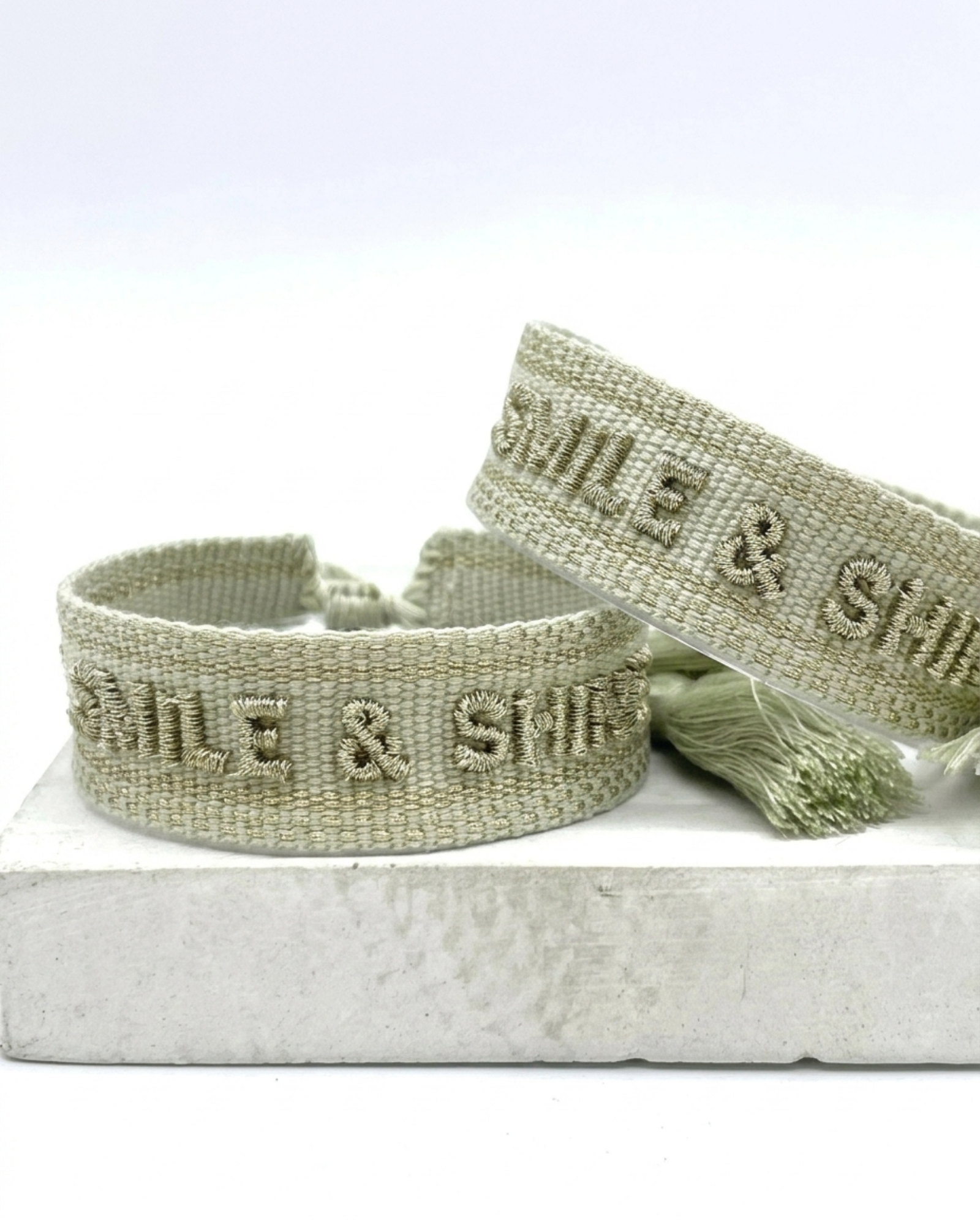 Close-up of adjustable platinum sage woven bracelet with tone-on-tone embroidered “SMILE & SHINE” lettering and sliding knot tassel cords.