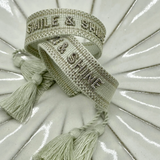 Close-up of adjustable platinum sage woven bracelet with tone-on-tone embroidered “SMILE & SHINE” lettering and sliding knot tassel cords.
