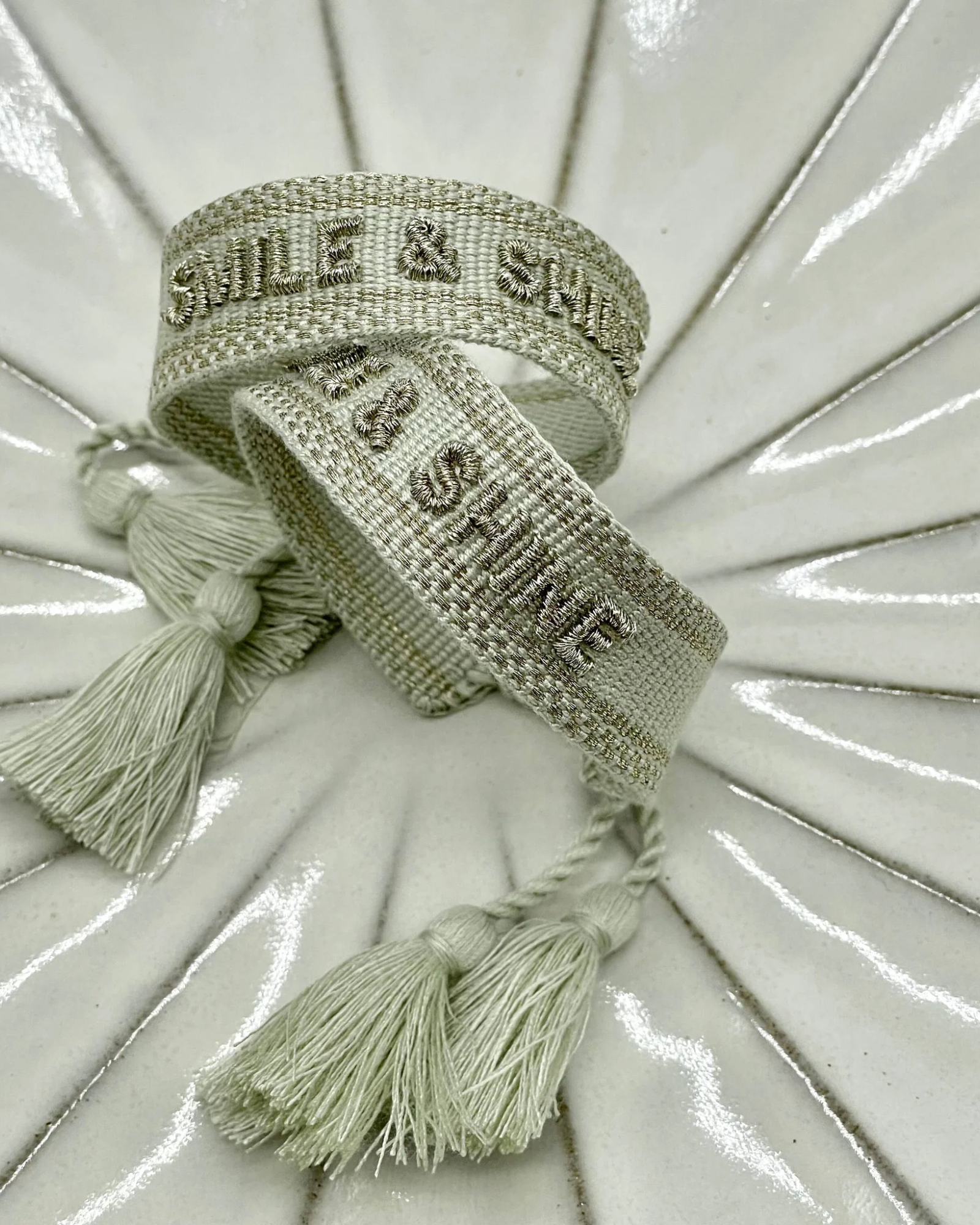 Close-up of adjustable platinum sage woven bracelet with tone-on-tone embroidered “SMILE & SHINE” lettering and sliding knot tassel cords.