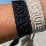 Close-up of adjustable dark grey woven bracelet with black embroidered “WHATEVER” lettering, black border, and sliding knot tassel cords.