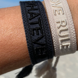 Close-up of adjustable dark grey woven bracelet with black embroidered “WHATEVER” lettering, black border, and sliding knot tassel cords.