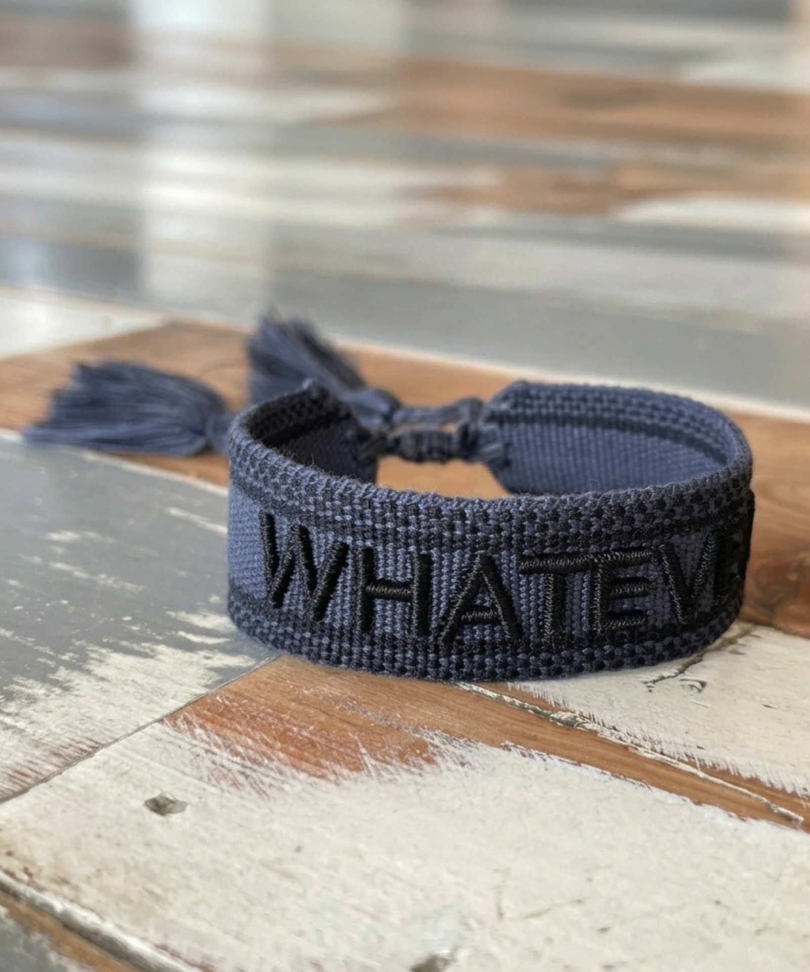 Close-up of adjustable dark grey woven bracelet with black embroidered “WHATEVER” lettering, black border, and sliding knot tassel cords.