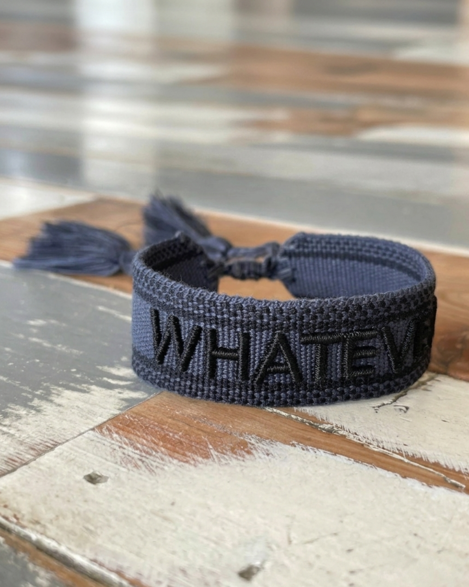 Close-up of adjustable dark grey woven bracelet with black embroidered “WHATEVER” lettering, black border, and sliding knot tassel cords.