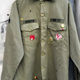 Front studio view of the khaki military shirt jacket hanging, showcasing the two chest pockets, full button closure, and extensive collection of pins and colorful custom patches.