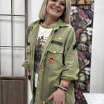 A smiling woman wearing the khaki green Rita military patch shirt jacket over a graphic t-shirt, displaying its straight fit and unique front patch details.