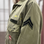 Close-up side view of the Rita military jacket, highlighting the classic khaki fabric texture and the black chevron patch embroidered on the sleeve.
