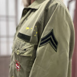 Close-up side view of the Rita military jacket, highlighting the classic khaki fabric texture and the black chevron patch embroidered on the sleeve.