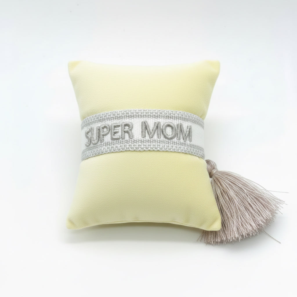 Silver embroidered Super Mom woven bracelet with blush tassel, displayed on a beige gift cushion — thoughtful Mother's Day bracelet gift