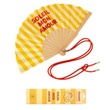 An opened summer folding fan with a vibrant yellow and white striped pattern and "SOLEIL MON AMOUR" text, displayed alongside its matching red shoulder strap and designer gift box.