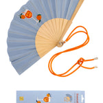 An opened summer folding fan with a soft blue fabric and a vibrant Spritz cocktail print, displayed alongside its contrast orange shoulder strap and matching packaging.