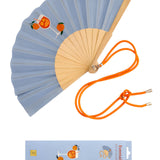 An opened summer folding fan with a soft blue fabric and a vibrant Spritz cocktail print, displayed alongside its contrast orange shoulder strap and matching packaging.