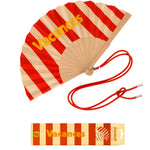 An opened summer folding fan with a red and beige striped pattern and bold yellow VACANCES typography, displayed next to its red shoulder cord and matching packaging.