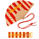 An opened summer folding fan with a red and beige striped pattern and bold yellow VACANCES typography, displayed next to its red shoulder cord and matching packaging.
