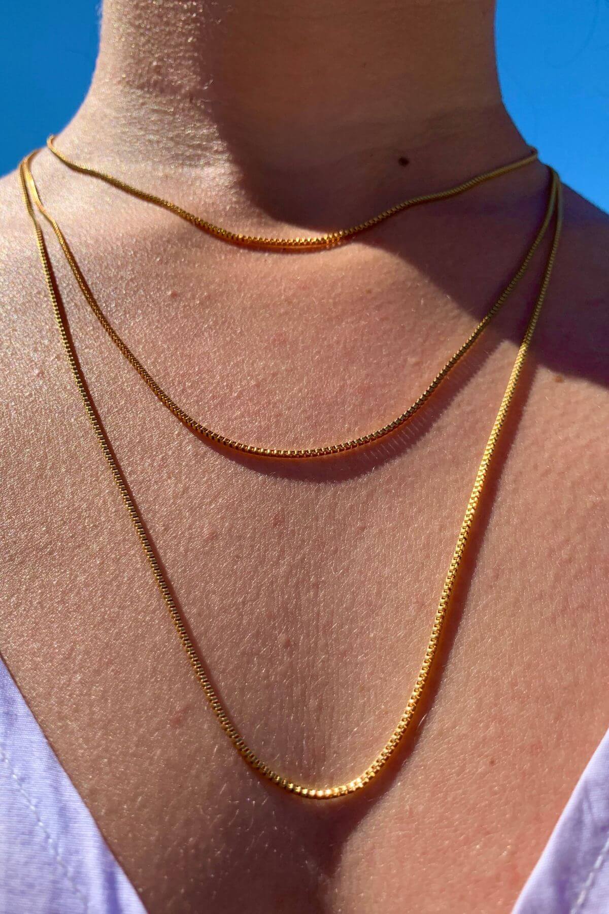 Crystal Haze gold-plated box chain necklace worn layered against a blue sky backdrop